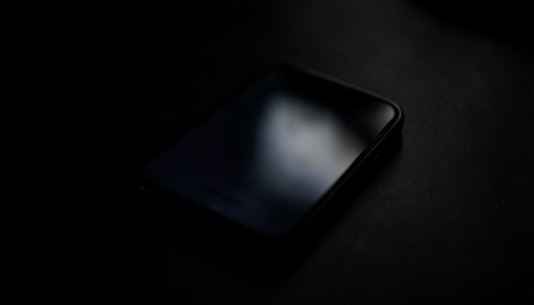 Dark close-up of a smartphone screen reflecting faint ambient light on a matte black surface, deep charcoal tones, low-key mood