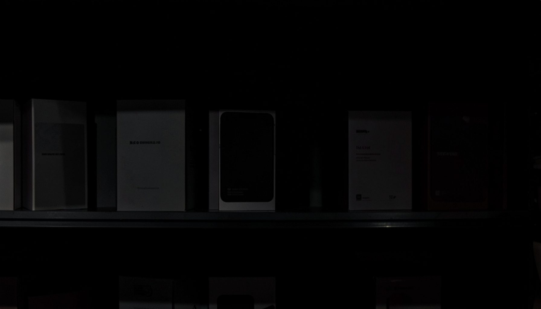Darkened retail display shelf with smartphone boxes in shadow, muted grays and deep blacks, subdued atmosphere